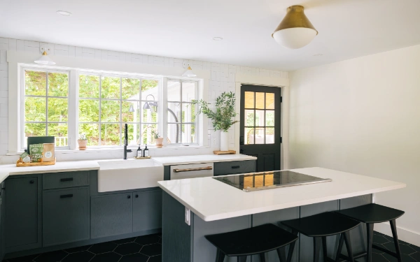 Bright kitchen with farmhouse sink, deep green cabinetry, white countertops, and large windows overlooking leafy outdoor views